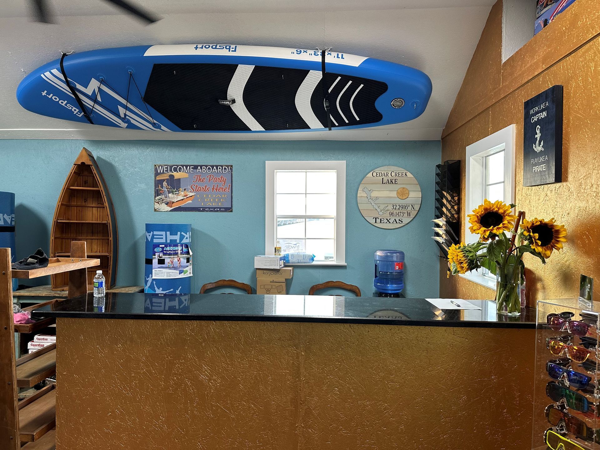 A surf-themed shop interior featuring a surfboard mounted on the ceiling, a countertop, and a vase of sunflowers.