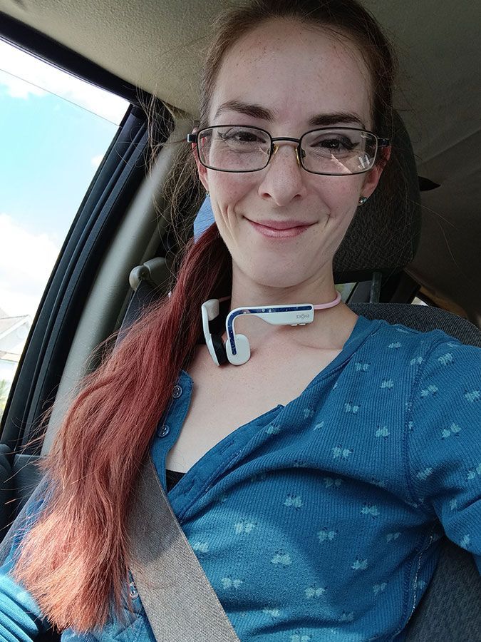 Woman in glasses and blue top smiling in a car, with a seat belt on and a small device around her neck