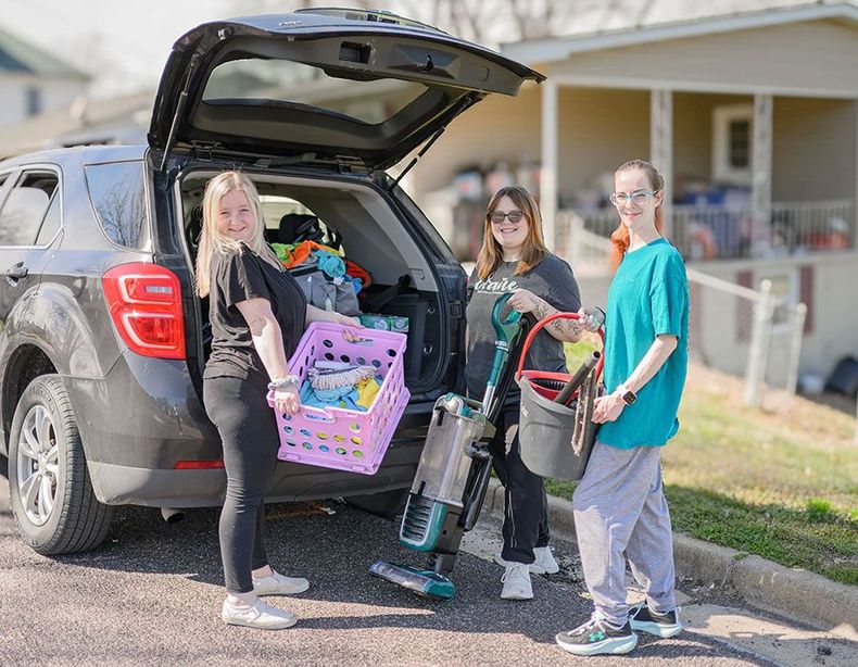 Three people unloading groceries and supplies from a black SUV in a driveway