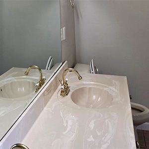 Bathroom sink with mirror, white countertop, brass faucet, and a toilet visible in the background
