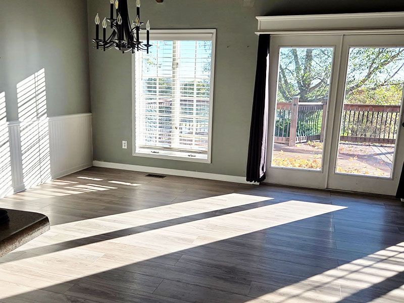 Empty living room with dark wood floors, chandelier, and sunlight through windows and glass doors.