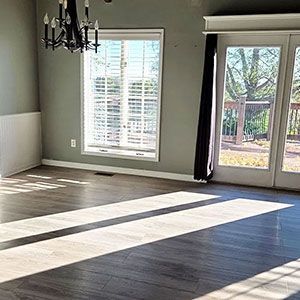 Empty living room with dark wood floors, large windows, and bright sunlight streaming in.