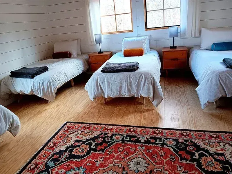 A bright bedroom with four single beds, wooden nightstands, and a red patterned rug on hardwood floors.