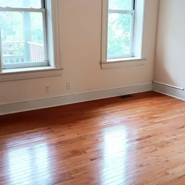 Empty room with polished hardwood floor, beige walls, and two windows with daylight.