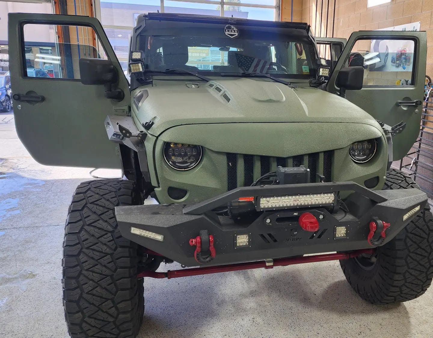 Olive-green off-road Jeep with open doors and rugged front bumper in a garage