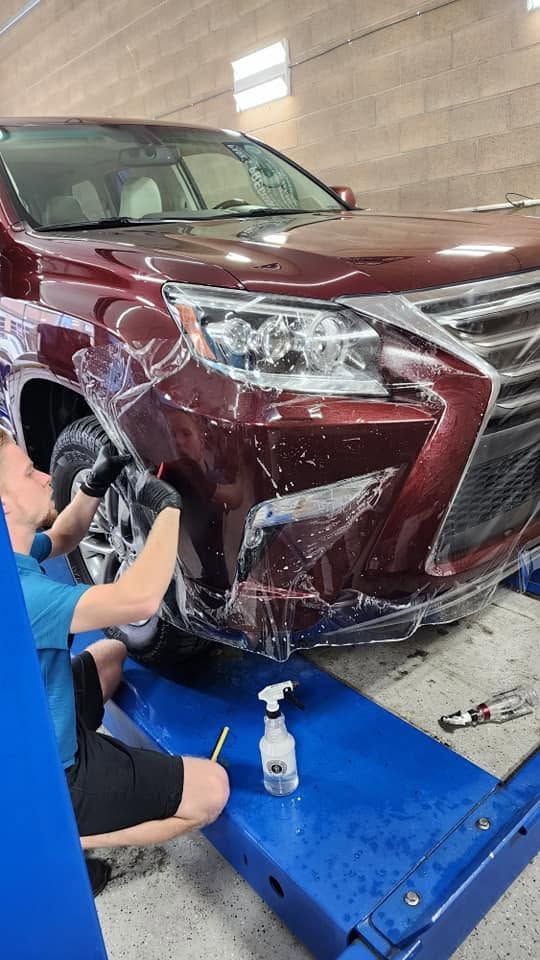 Mechanic cleaning a maroon SUV’s front bumper in a workshop, with a pressure washer and foam.