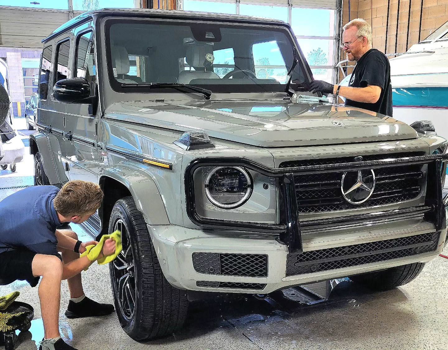 Two men detail a silver Mercedes G-Class SUV indoors, one polishing the front wheel and bumper.