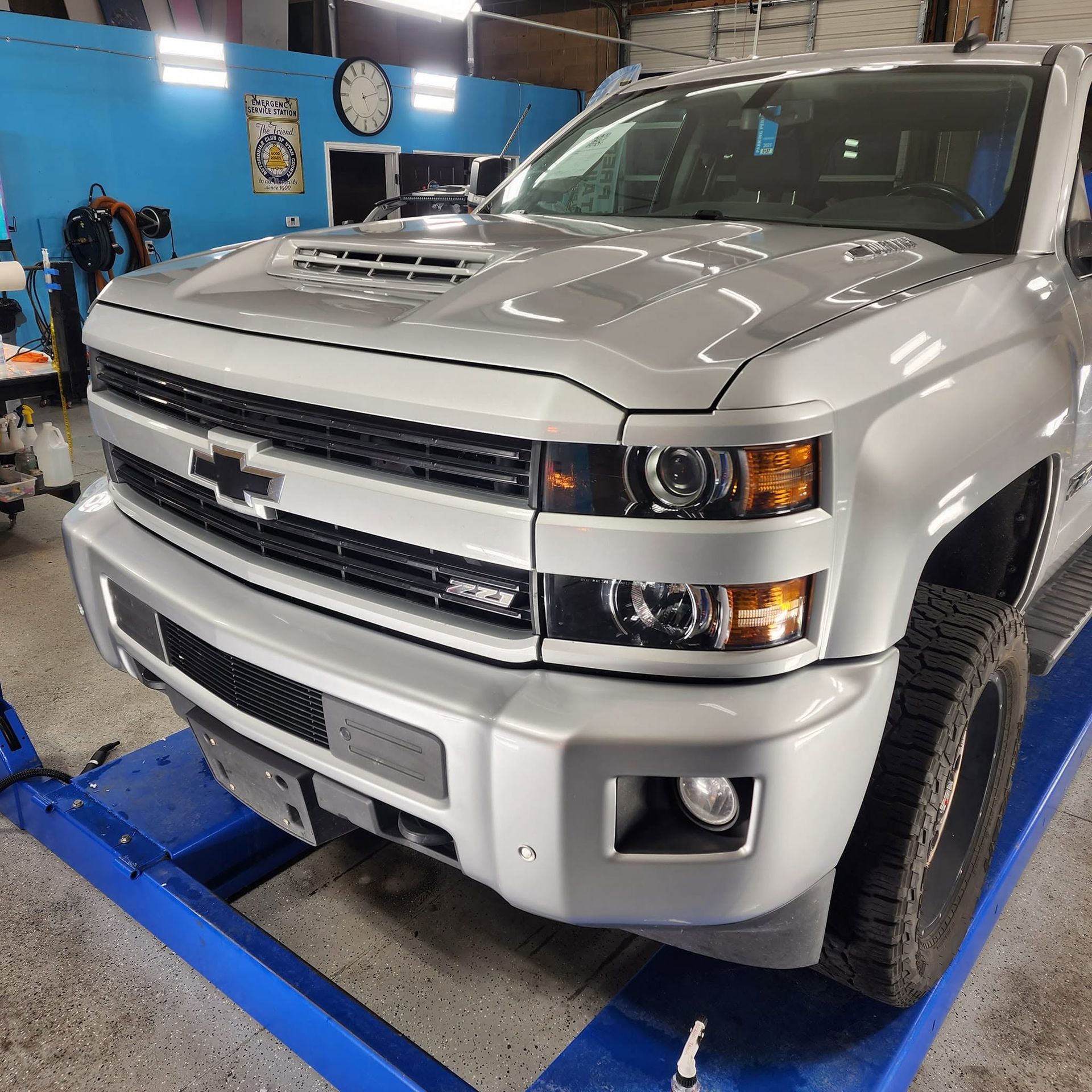 Silver Chevrolet pickup on a lift in an auto shop, front view with hood vents and blue lift arms