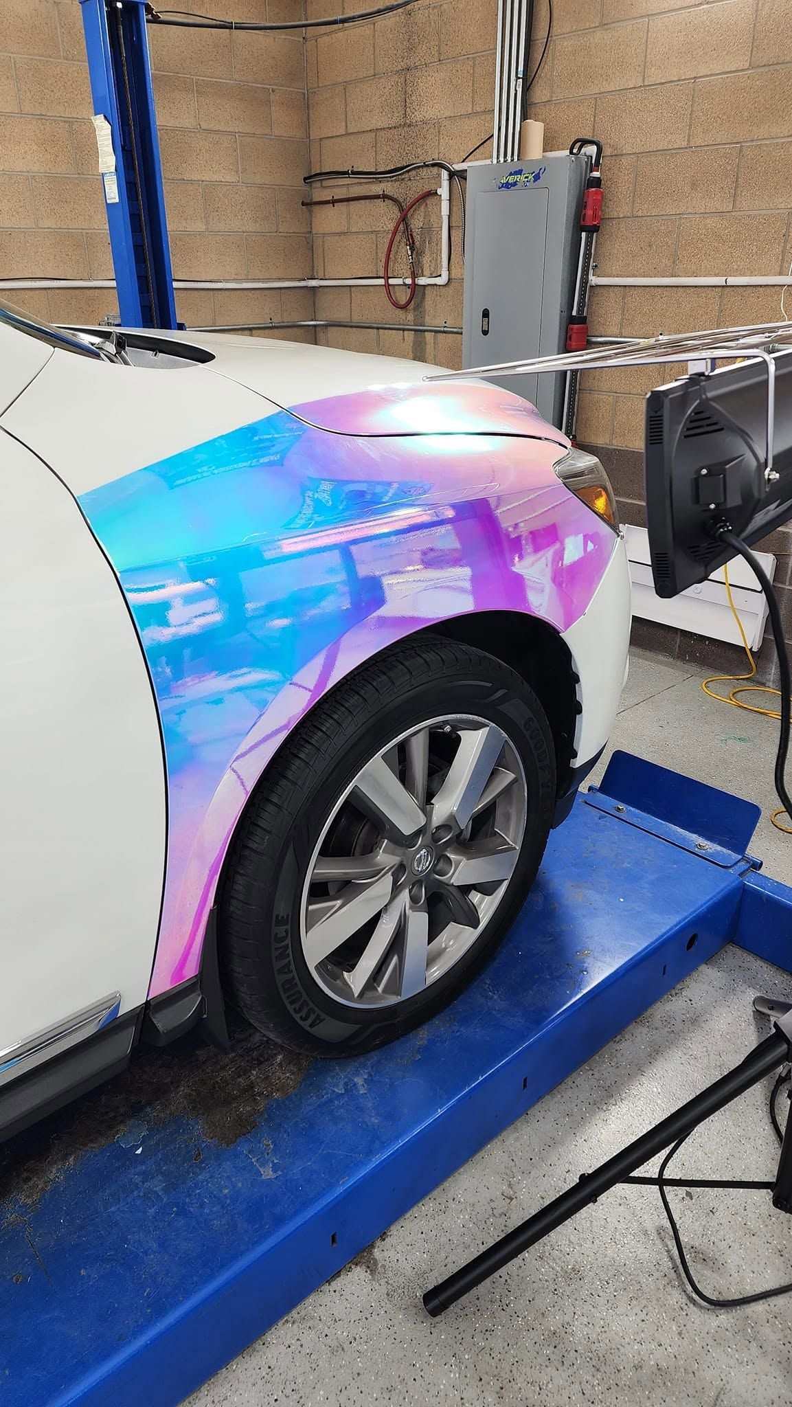 Electric car on a lift in a garage, with iridescent wrap on the front fender.
