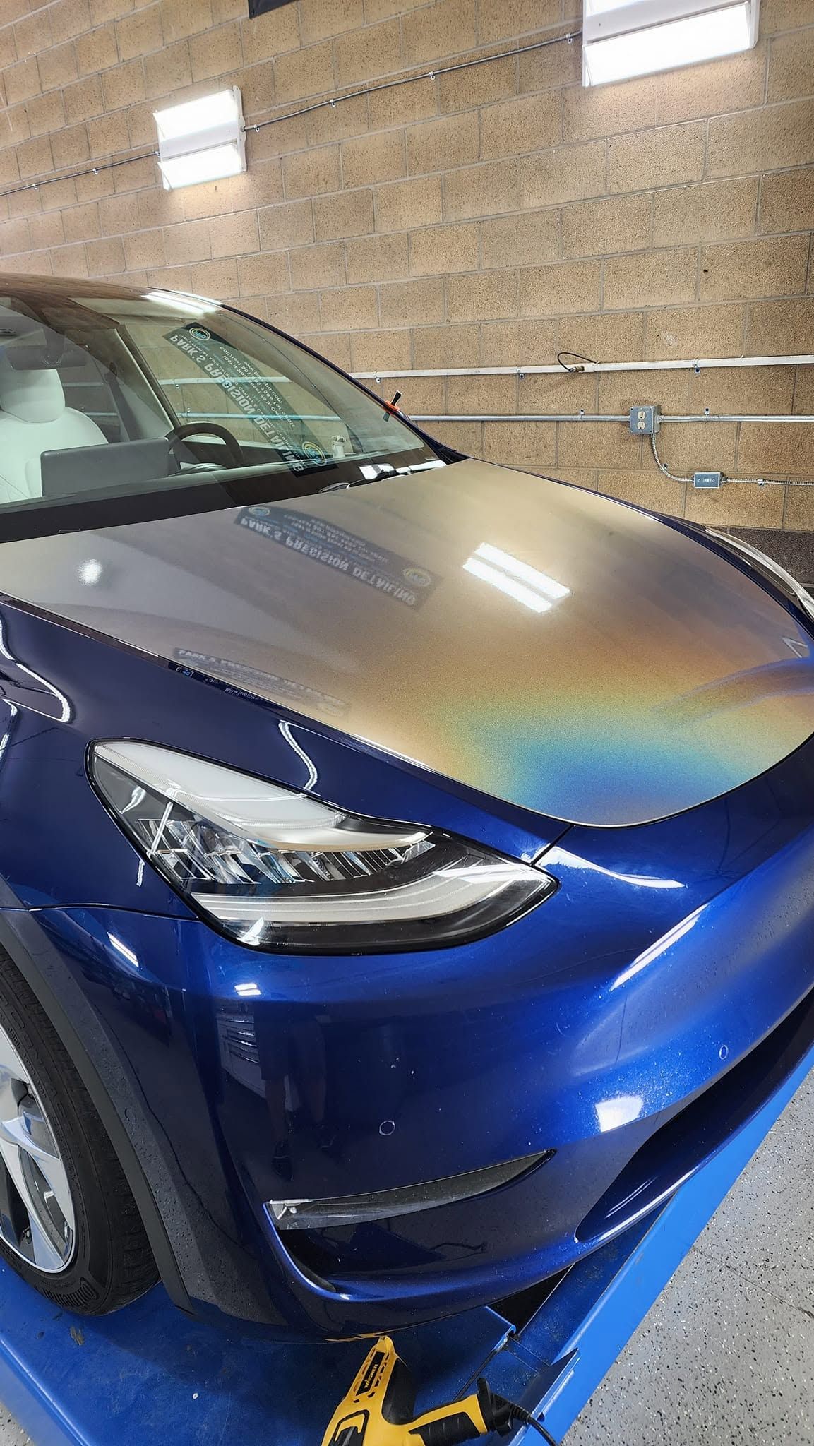Blue car parked indoors against a brick wall, front-left view with lights reflecting on the hood