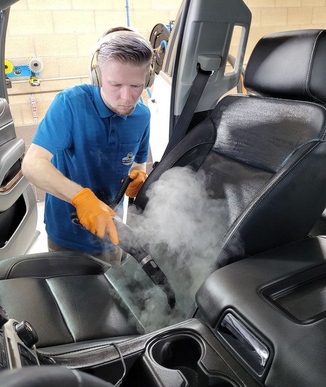 Detailing worker steam-cleans a black car seat interior with orange gloves.