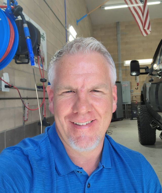 Smiling man in a blue shirt standing in a vehicle workshop with tools and an American flag in the background