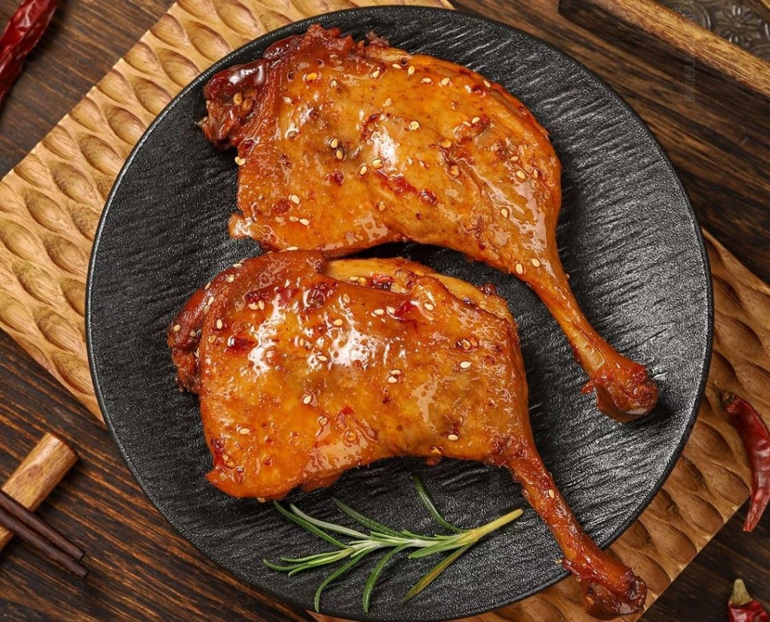Two glazed roasted chicken legs on a black plate with rosemary on a woven placemat.