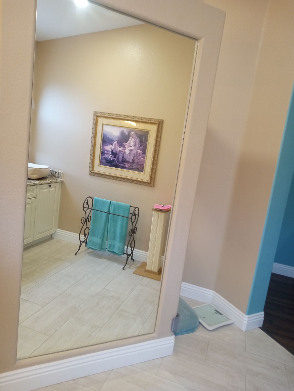 Large mirror reflecting a beige room with a framed picture, teal chair, and tiled floor.