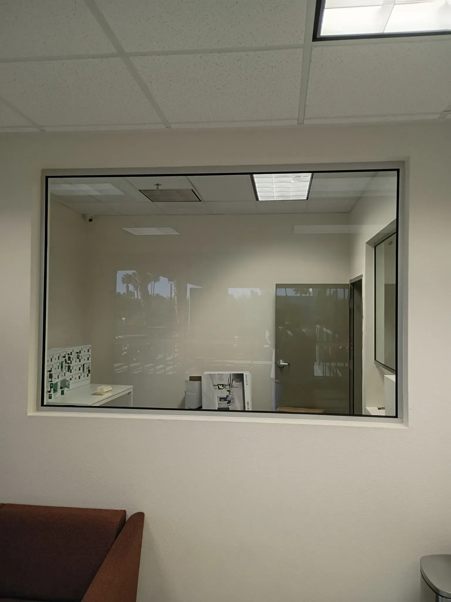 Office lobby with a large interior window looking into a quiet room and ceiling lights.