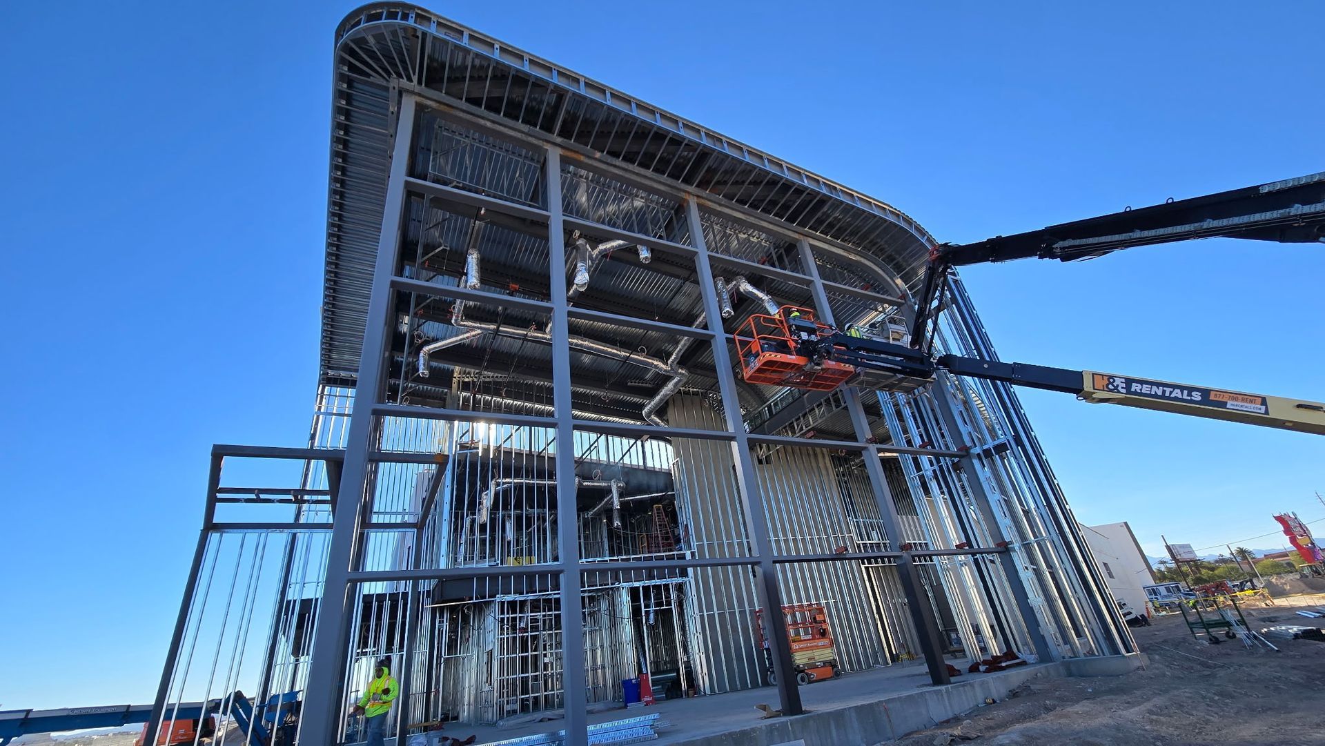 Large steel-framed building under construction with workers on a lift against a blue sky