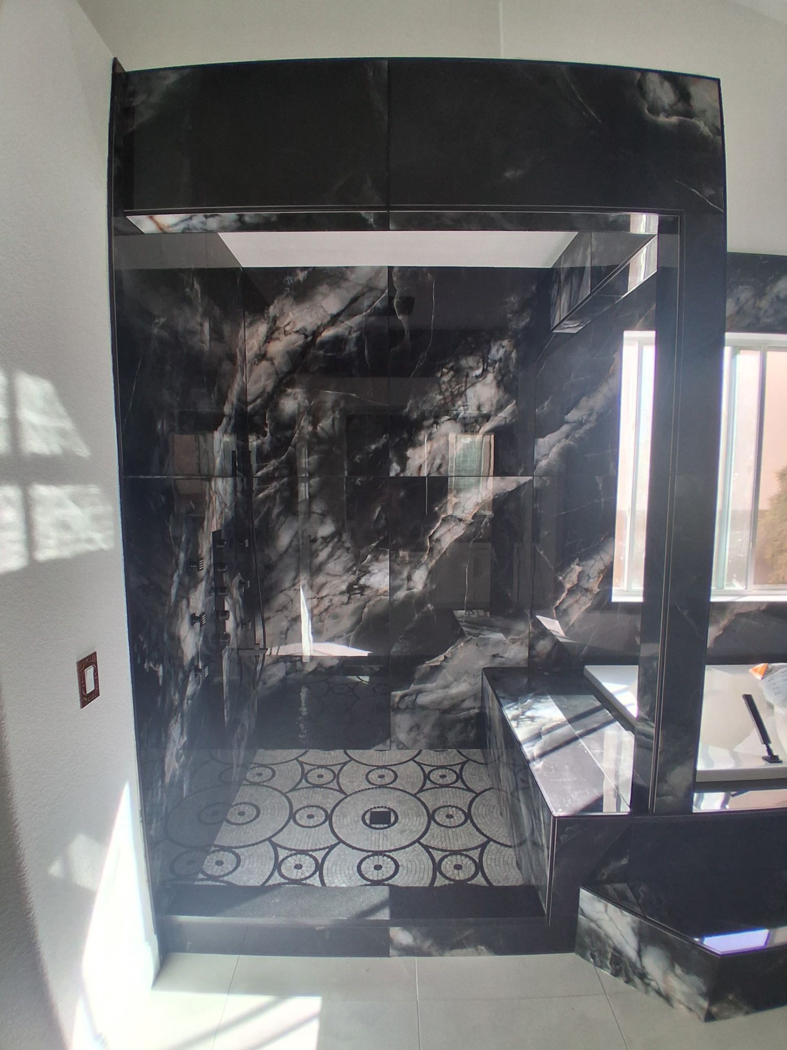 Black-tiled bathroom with a shower area, patterned floor, and a bright window on the right.