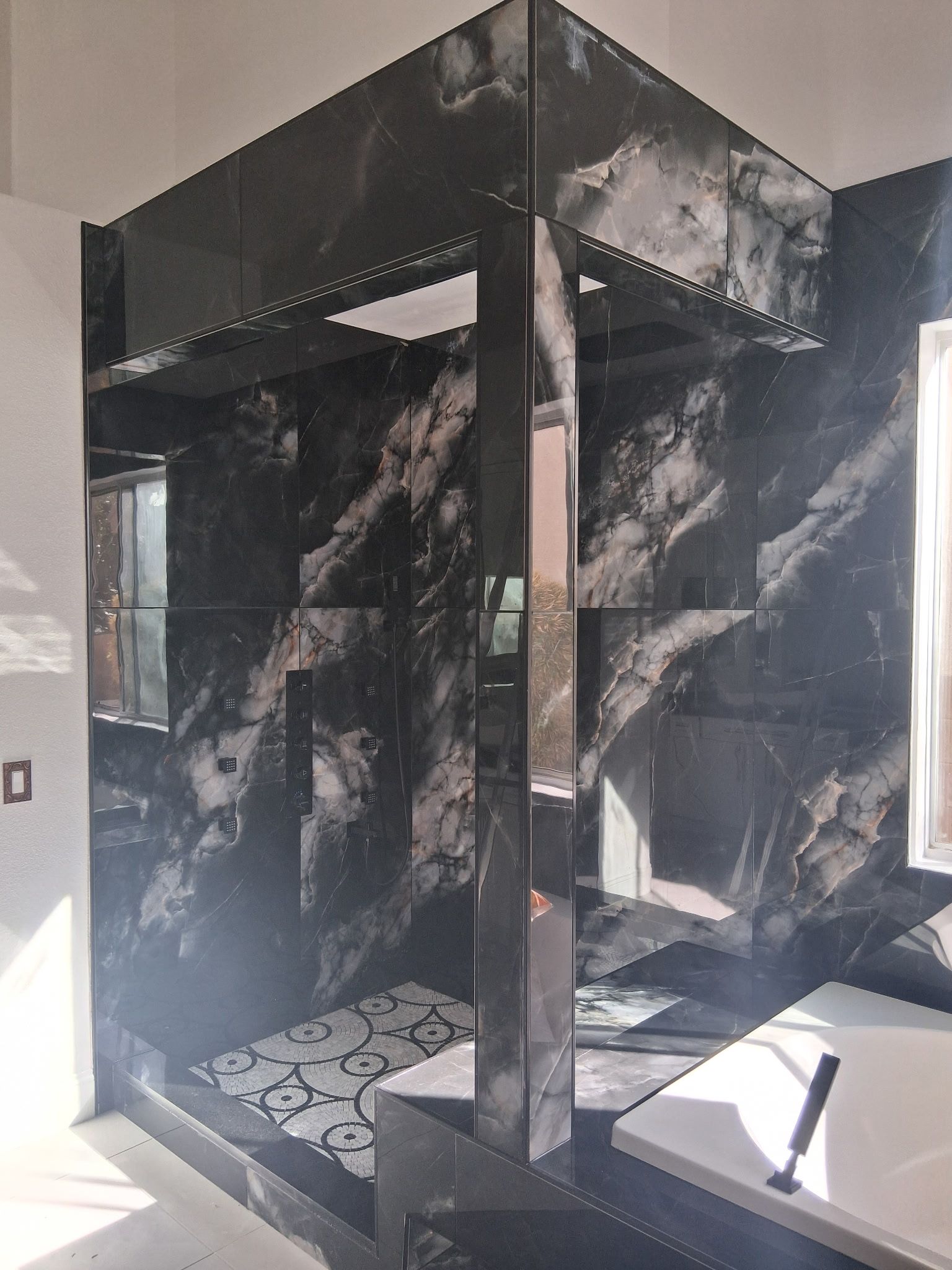 Glossy black marble shower stall with glass panel in a small bathroom, lit by sunlight