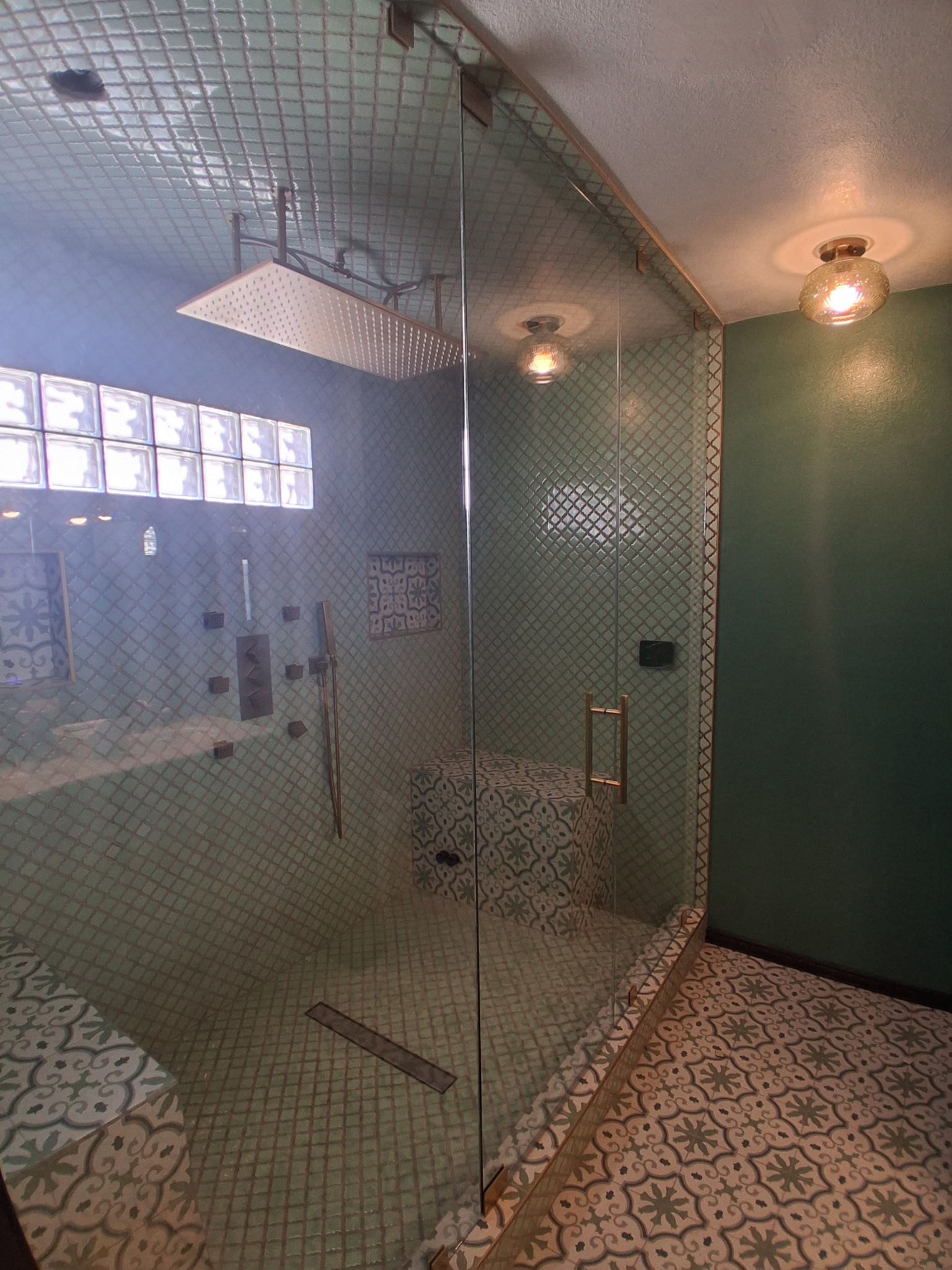 Glass shower enclosure in a tiled bathroom with patterned floor and wall sconces