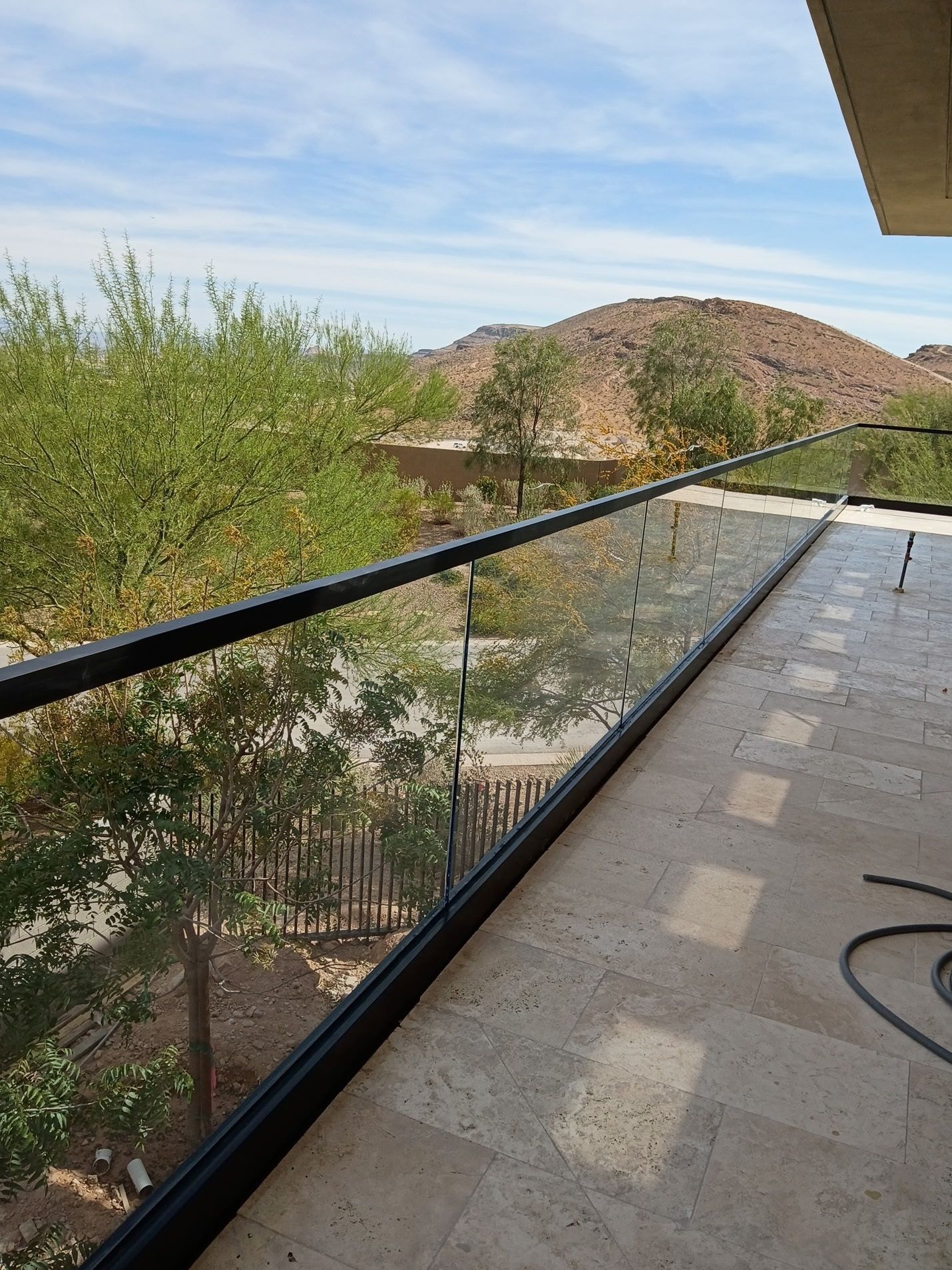 Balcony with glass railing overlooking trees, desert hills, and a paved terrace under a blue sky