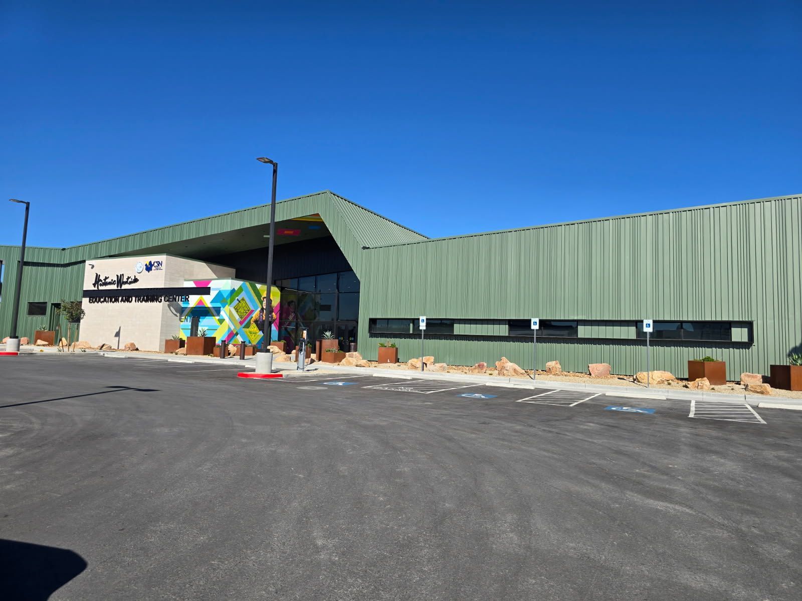 Green warehouse-style building with a mural, colorful sculpture at the entrance, and an empty asphalt parking lot.
