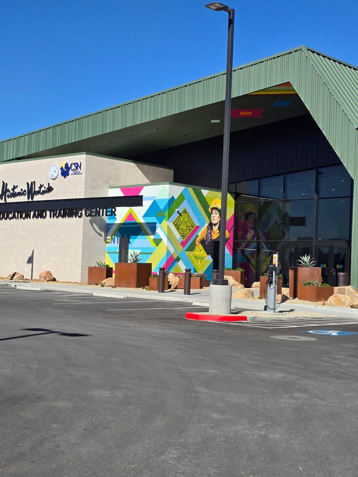 Modern building with colorful geometric mural and Arabic signage under a green roof, viewed from a parking lot
