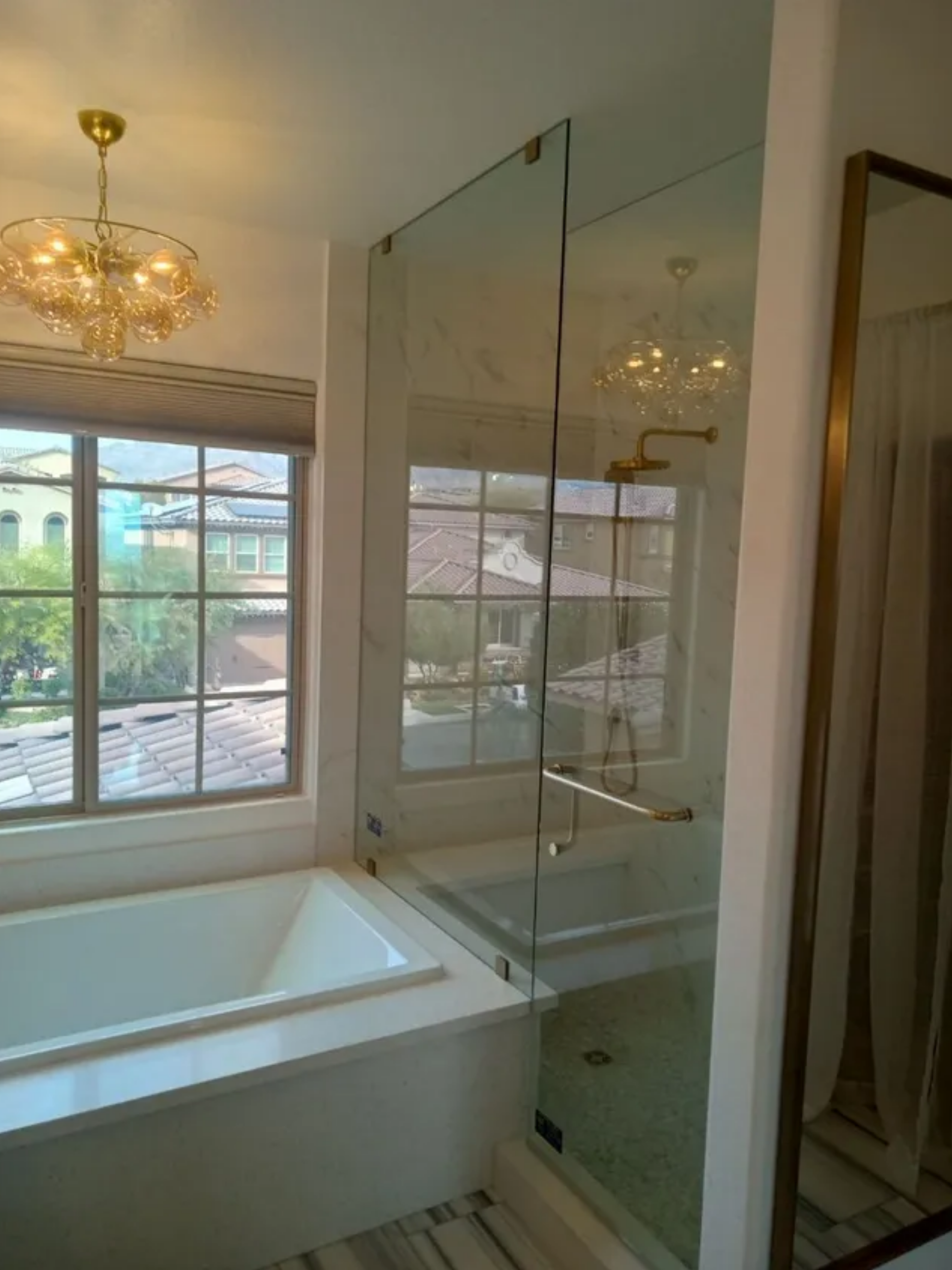 Bright bathroom with a soaking tub, glass shower enclosure, chandelier, and large window with a view outside.