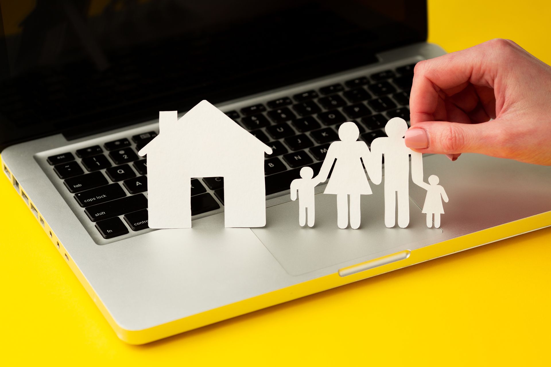 Paper cutout of a house and family on a laptop keyboard; hand holding family figure against a yellow background.