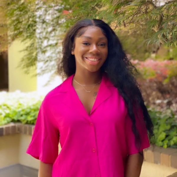Smiling person in a bright pink shirt standing outdoors by a garden walkway.
