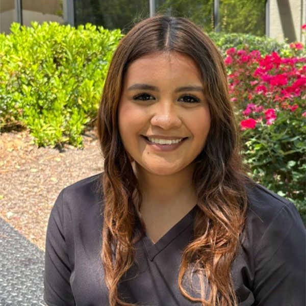 Smiling woman in dark gray scrubs standing outdoors with greenery and pink flowers behind her
