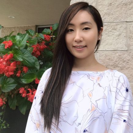 Woman standing outdoors beside red flowers and a stone wall, wearing a white patterned top and smiling gently
