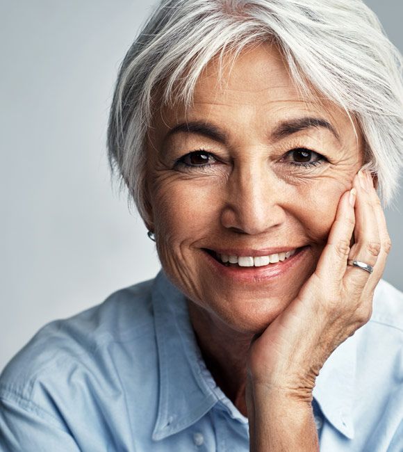 Smiling person with white hair resting a hand on their cheek, wearing a light blue shirt.