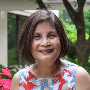 Smiling woman in a red, white, and blue floral top standing outdoors near greenery and pink flowers