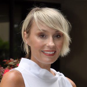 Blonde woman smiling in a white sleeveless top with a high collar, standing outdoors.