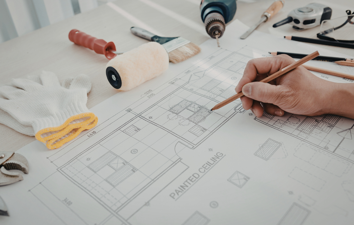 Hand drafting a floor plan with pencil beside blueprints, tools, and paint rollers on a worktable