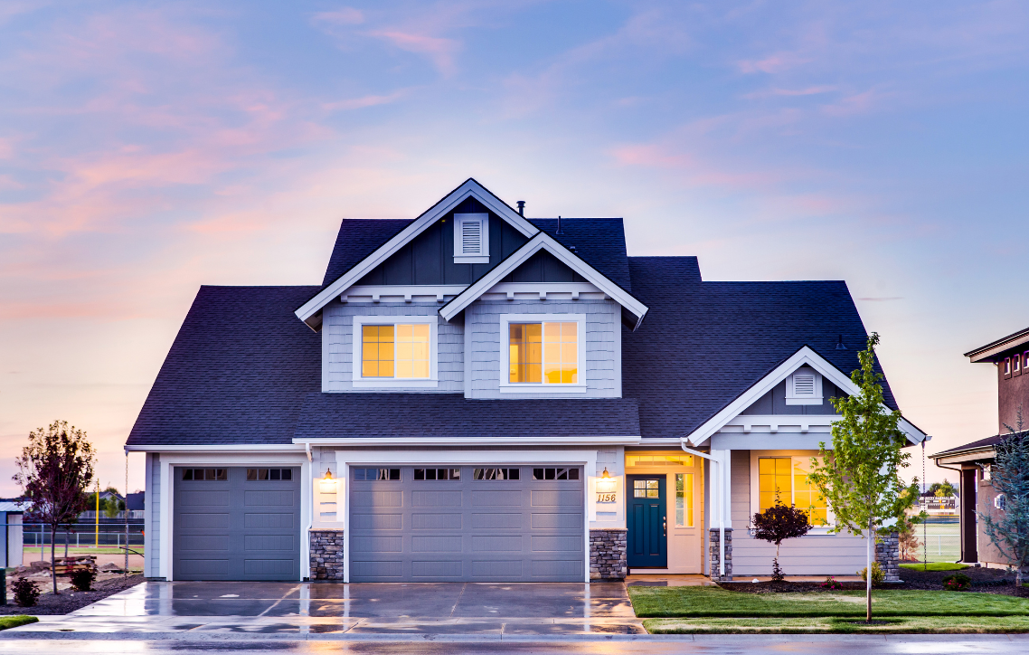 Modern gray two-story house with lit windows and a wet driveway at dusk
