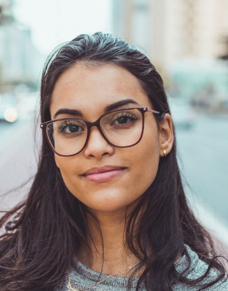 Portrait of a person with glasses and long dark hair, standing outdoors with a soft blurred city background
