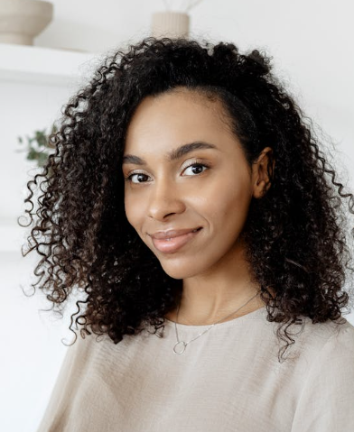 Portrait of a smiling person with curly hair in a light sweater, standing indoors against a bright background