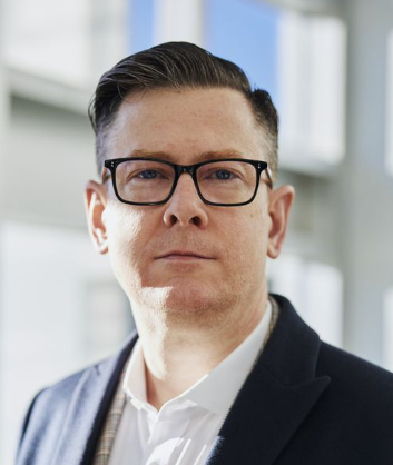 Portrait of a man in glasses and a dark blazer against a bright office background