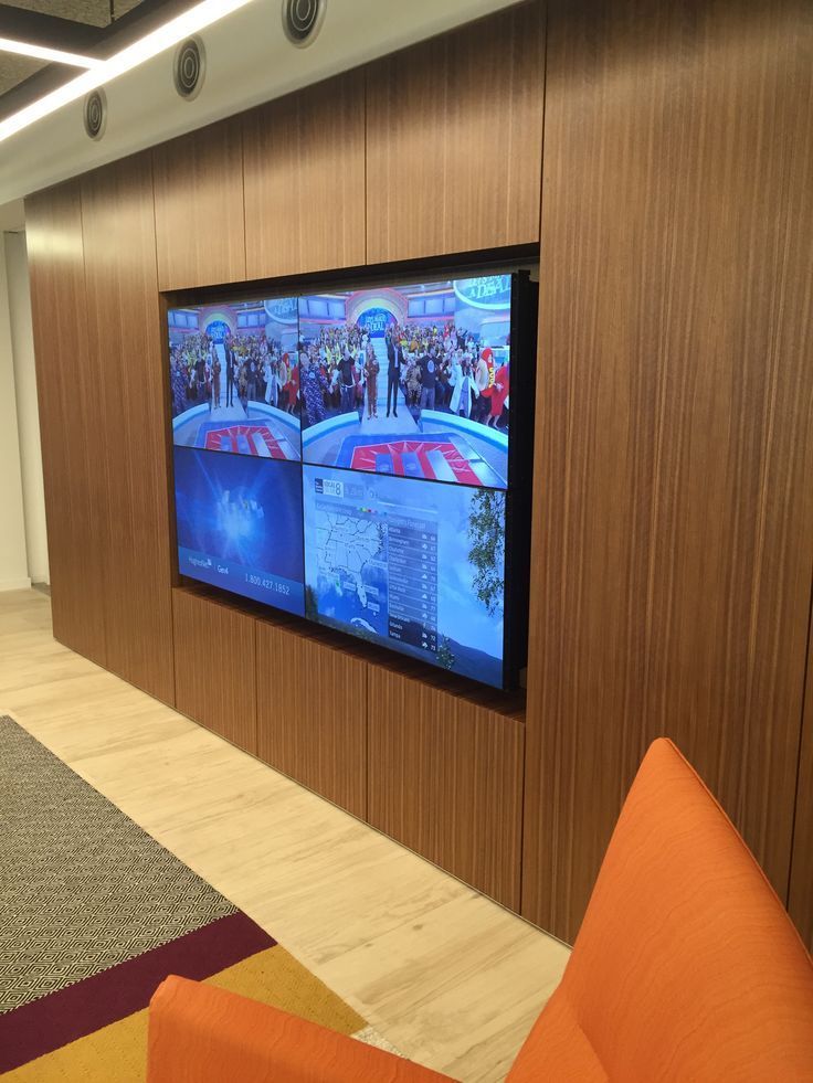 A wall-mounted TV showing a four-panel display, set against a wooden panel. An orange chair is in the foreground.