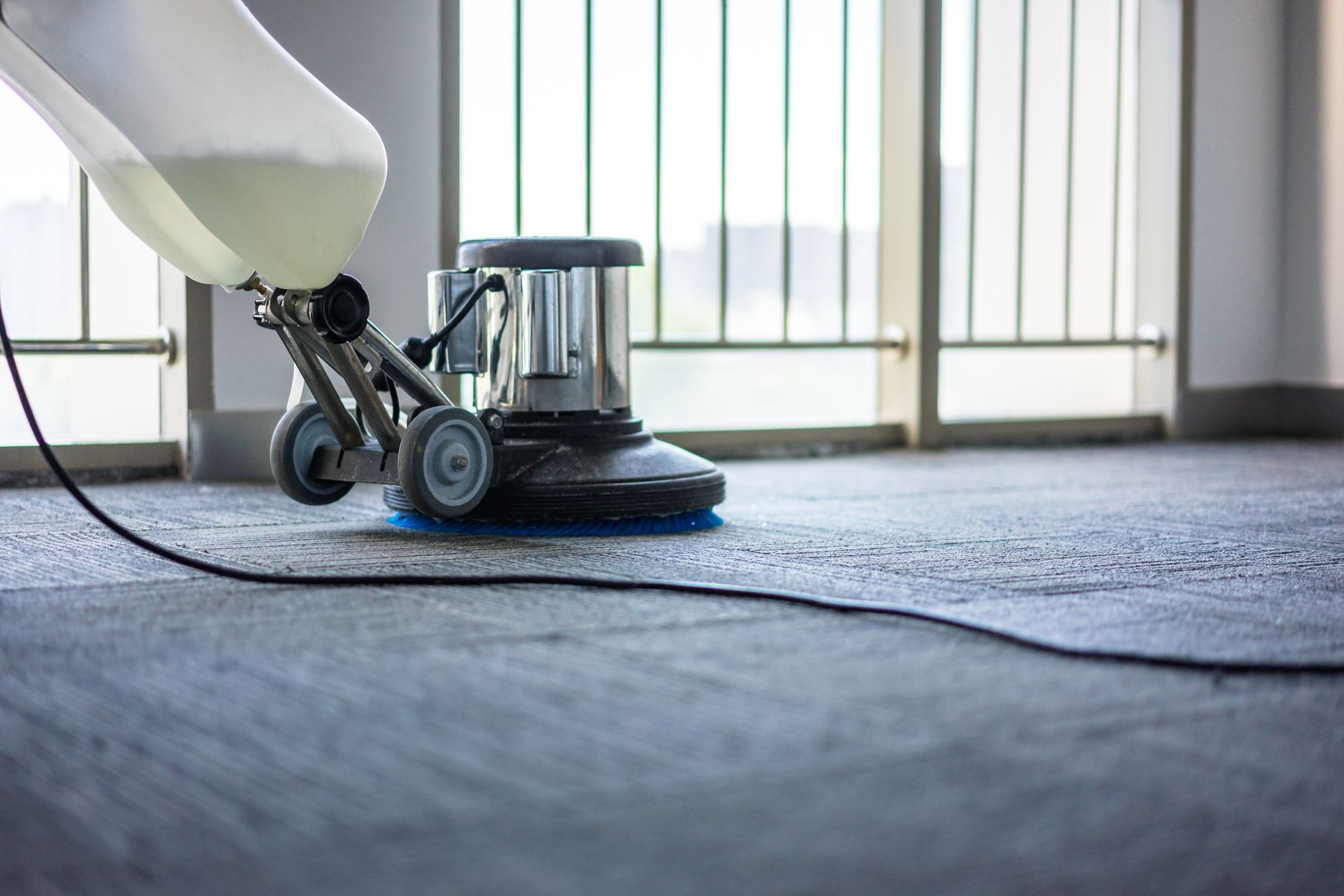 Floor cleaning machine applying cleaning solution to gray carpet near a window.