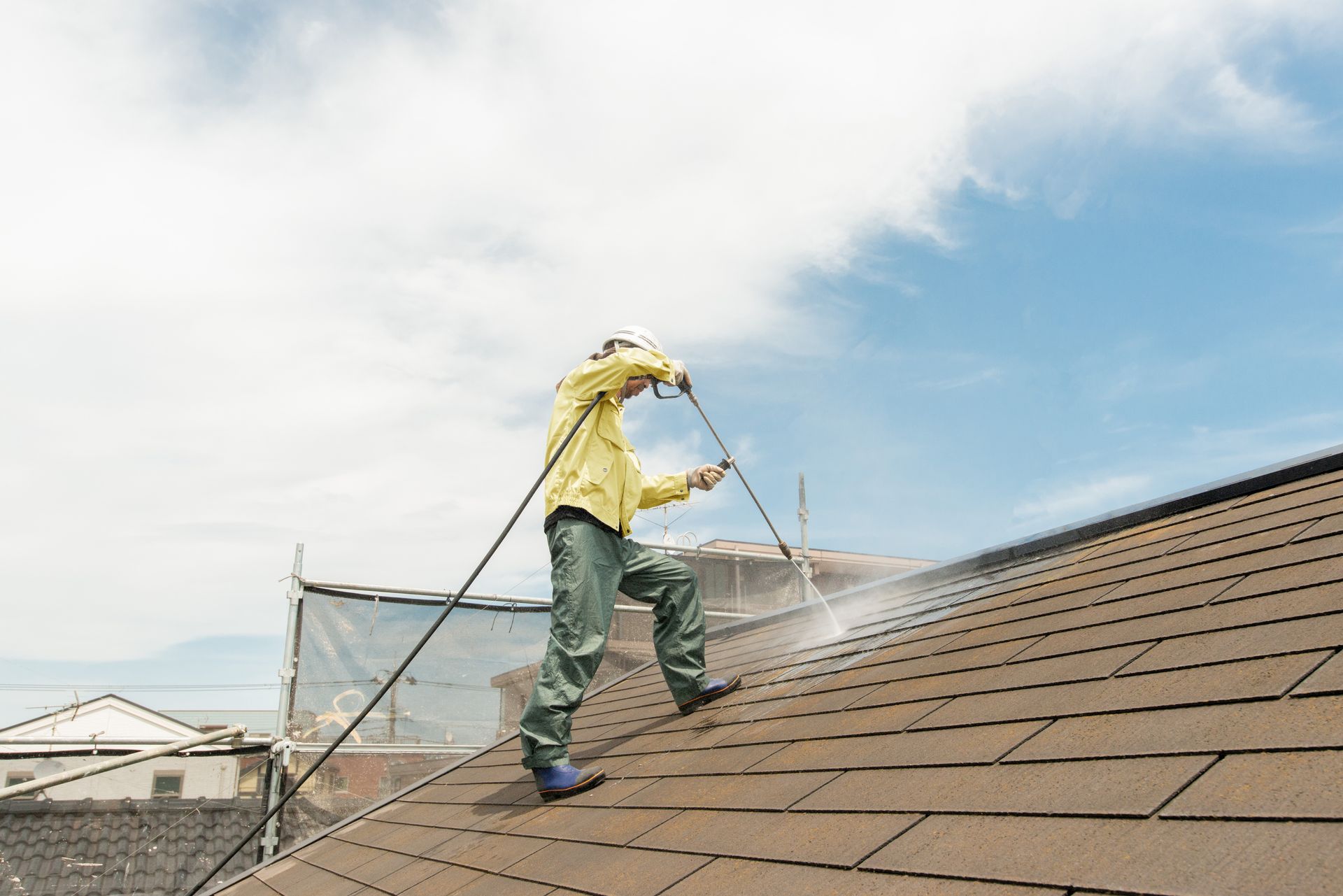 Man in protective gear pressure washing a brown shingle roof on a sunny day.