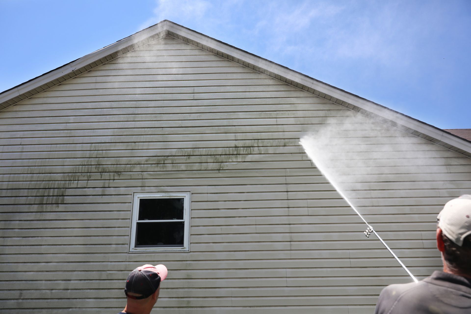 Man power washing a light-colored house siding to remove green algae. Blue sky in background.