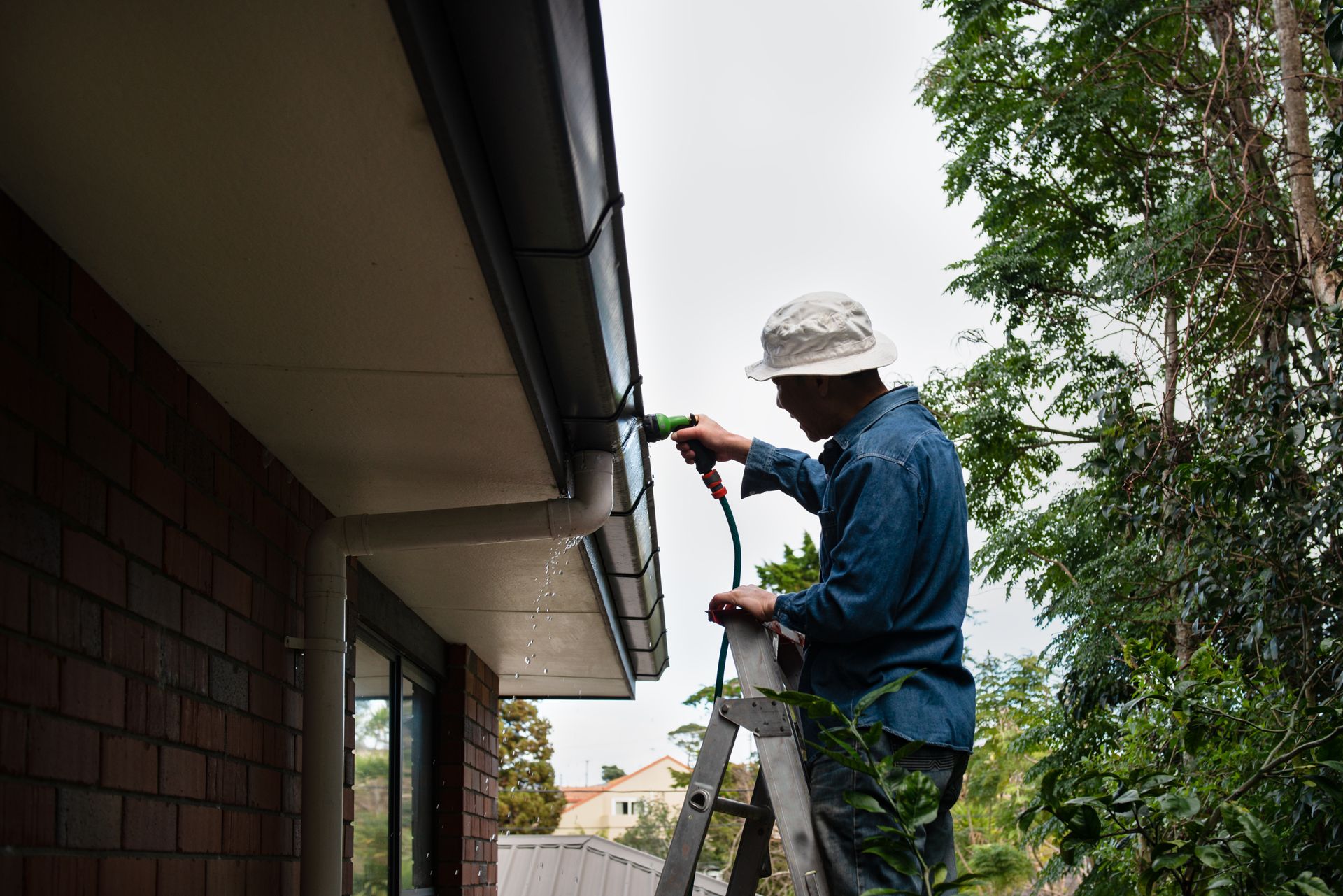 Person on a ladder cleaning a rain gutter on a brick building, using a tool, surrounded by trees.
