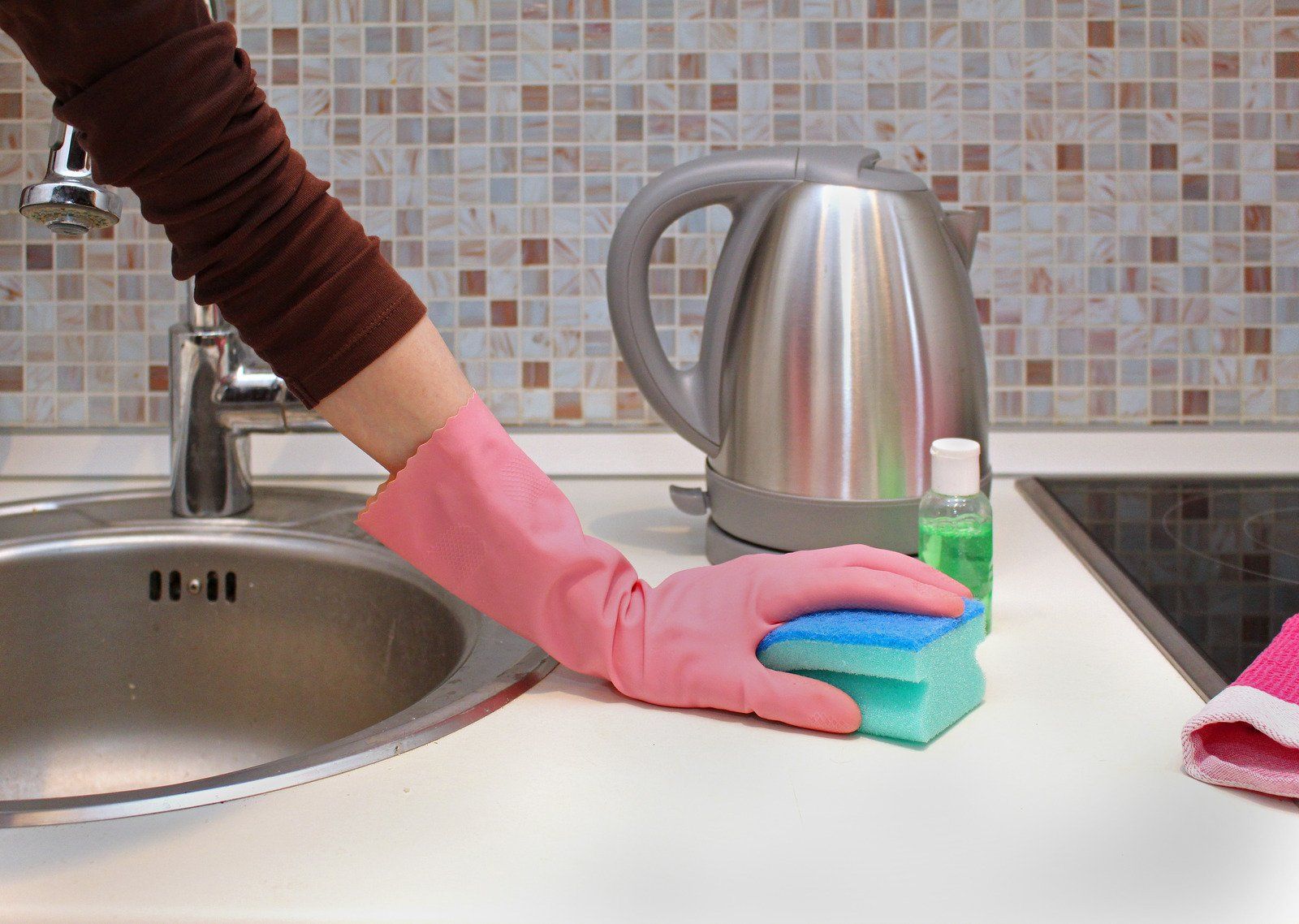 Person in pink glove cleans white countertop with a sponge near sink and electric kettle.