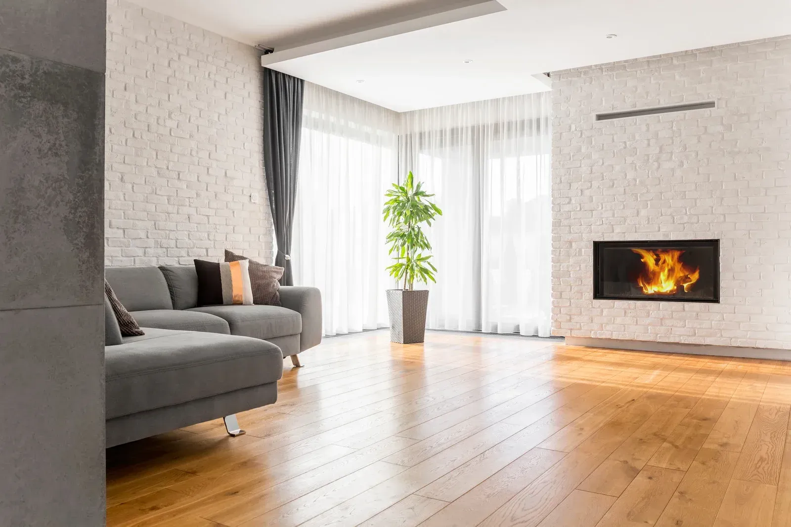Cozy living room with gray sofa, fireplace, and hardwood floors. Large windows with sheer curtains.