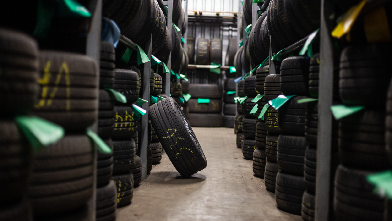 Stacks of black tires lined in an industrial aisle, with green tags and one tire leaning in the center.