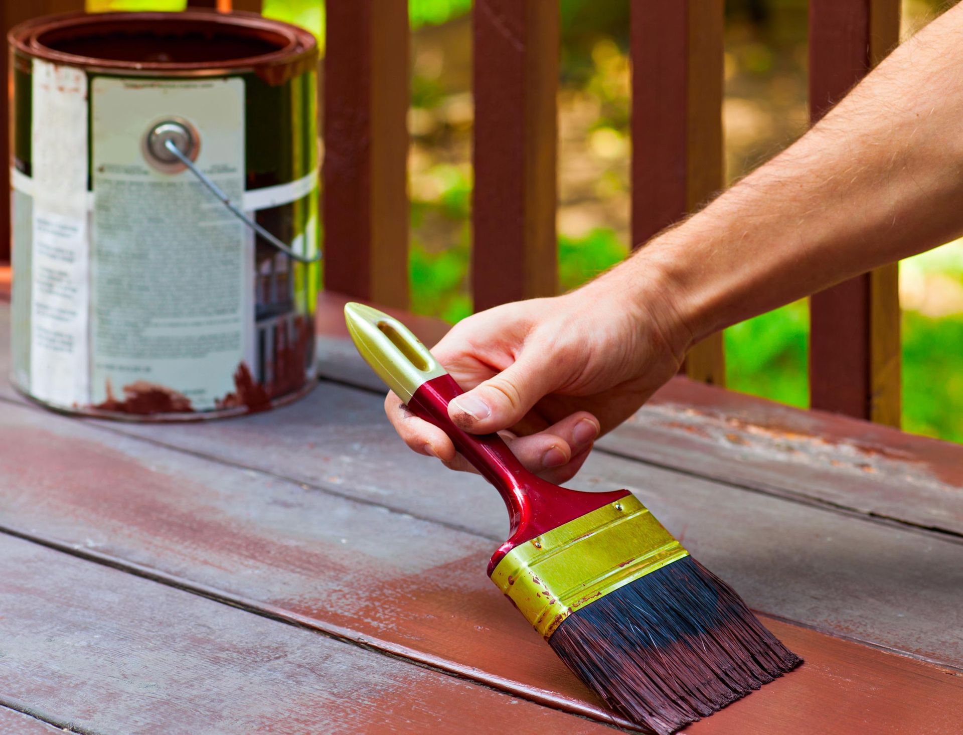 Person painting a wooden deck brown with a paintbrush and paint can nearby.