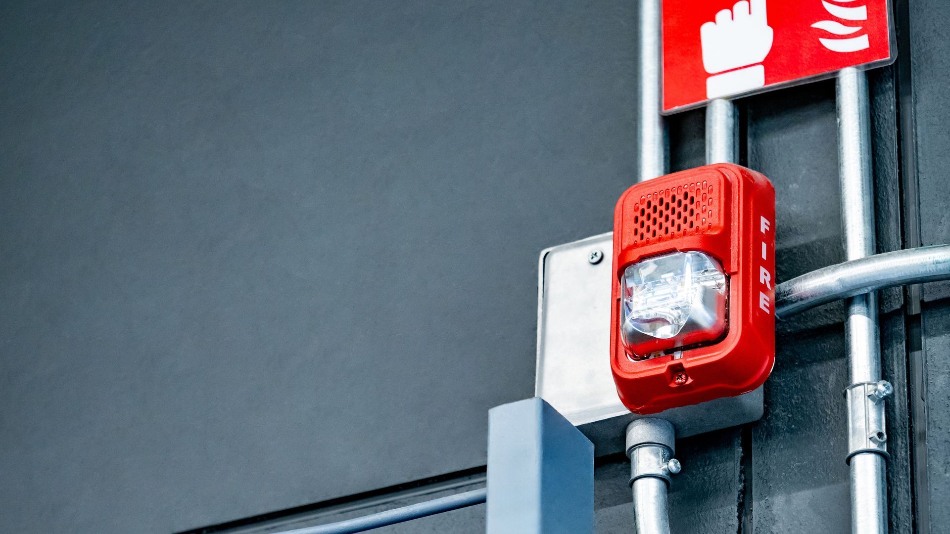 Red fire alarm with flashing strobe light, mounted on a gray wall with conduit pipes, and a red fire safety sign. Red fire alarm with flashing strobe light, mounted on a gray wall with conduit pipes, and a red fire safety sign.