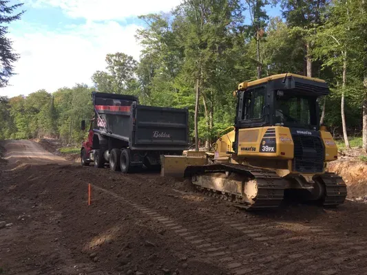 Un bulldozer nivelle un chemin de terre tandis qu'un camion-benne attend. Forêt en arrière-plan.